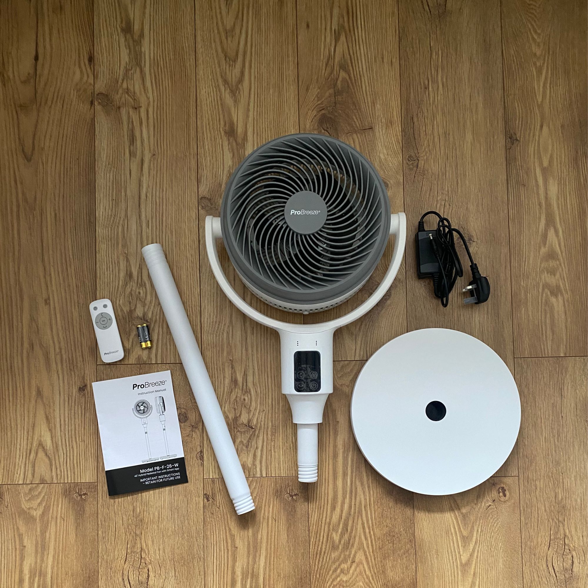 Flat lay of the parts that are included in the box when you buy the Pro Breeze Airflo 43'' Hybrid Pedestal Fan, including the fan head, base, pole, remote control, batteries, power cable and instruction manual on a wood-effect floor