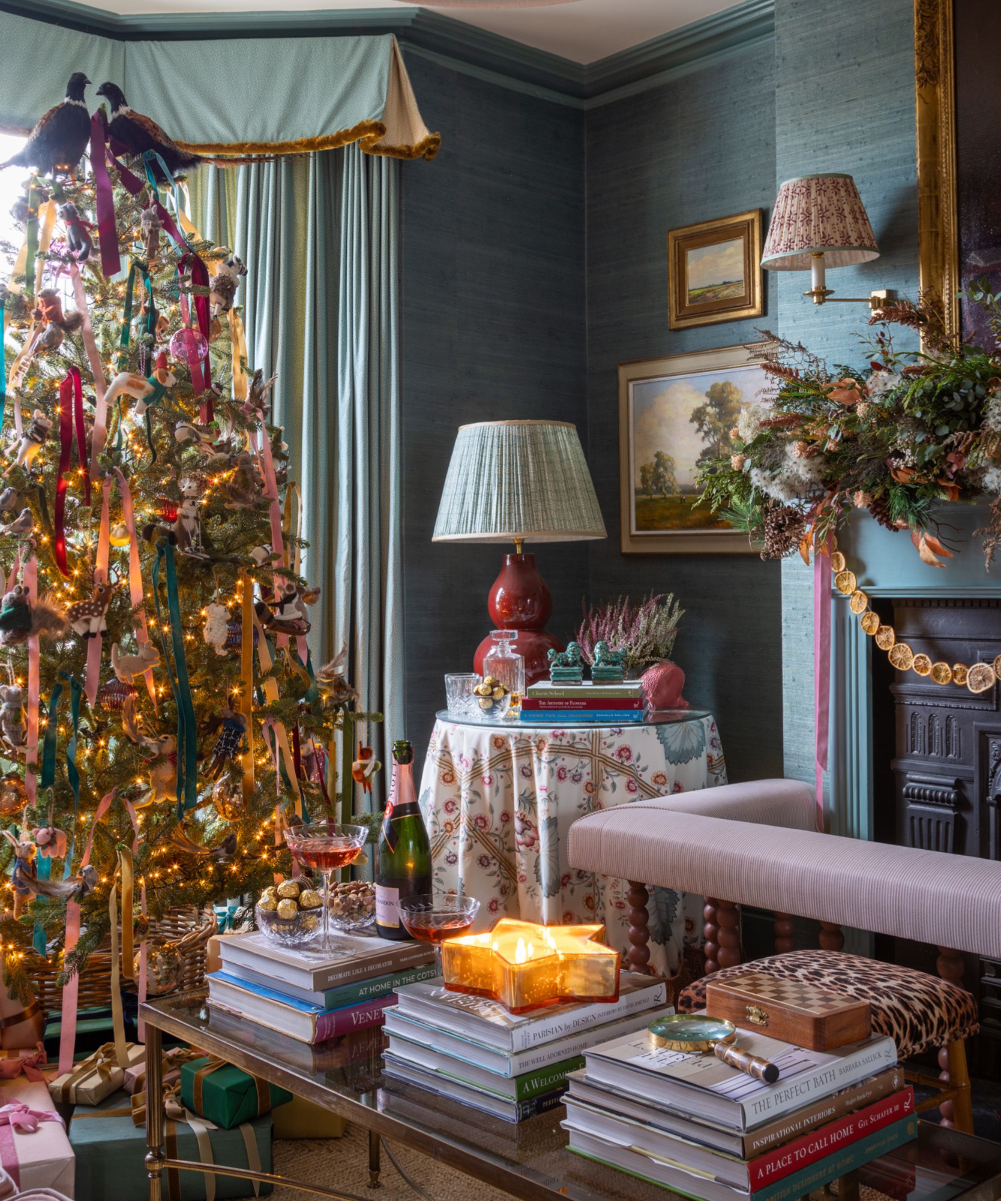 traditional english country living room wallpapered in blue grasscloth styled for christmas with a large velvet ribbon adorned tree topped with two faux pheasants and a fireplace styled with a large garland and dried oranges