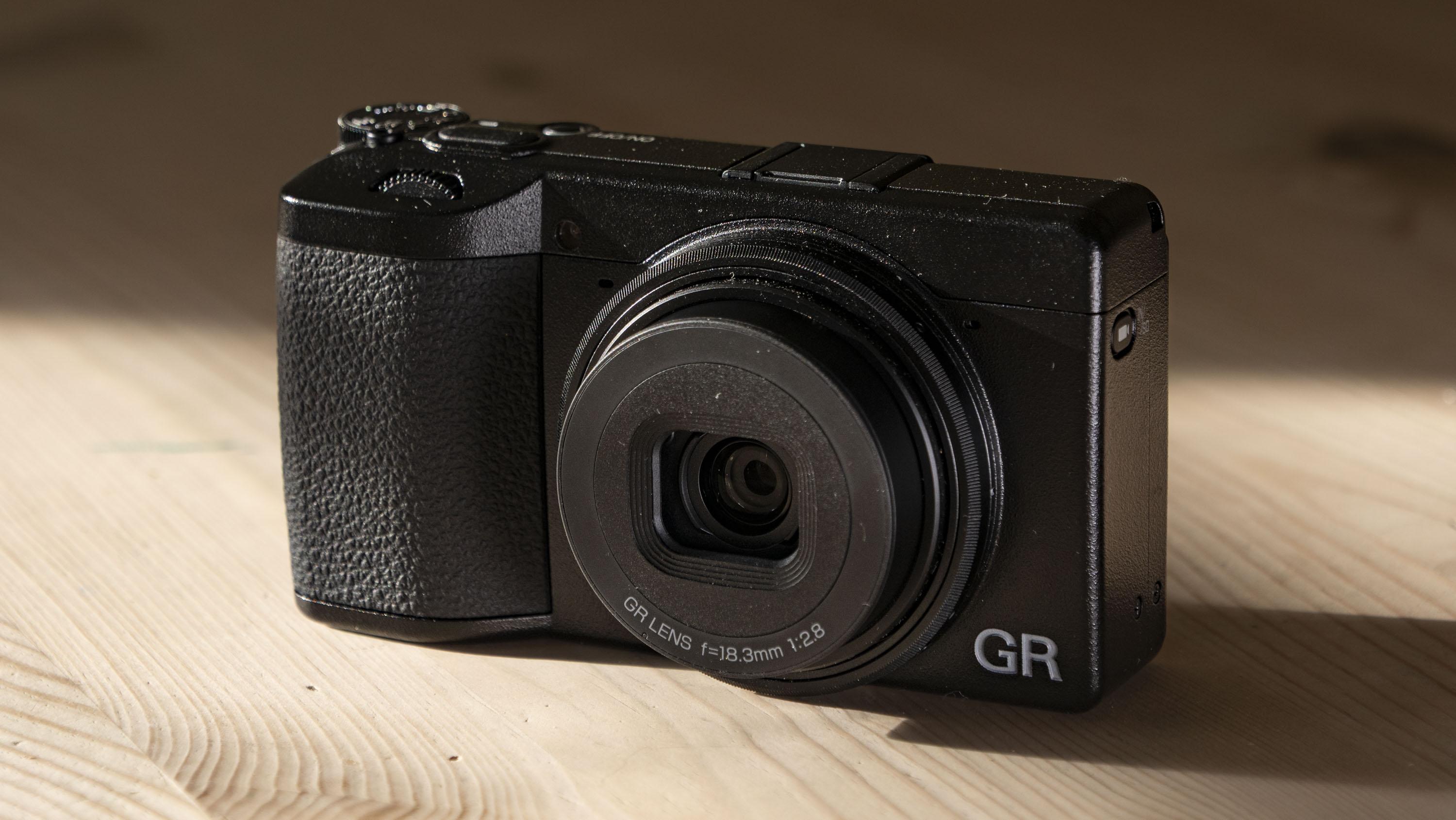 Ricoh GR IV compact camera on a wooden table, with strong sidelight and dark shadows