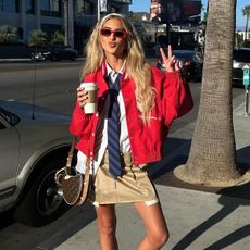 Woman wearing red jacket, white button down shirt, black tie, pleated mini skirt and sneakers.