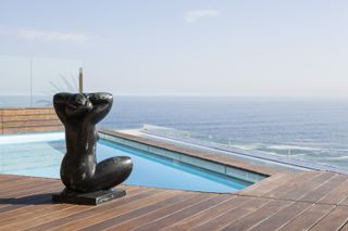 A pool with board decking and a black stone figure of a woman sitting with crossed legs and arms. It looks out onto the ocean.