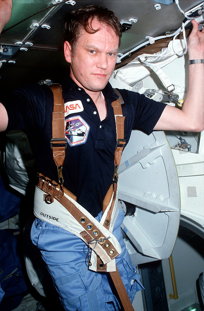a man in a collared blue nasa shirt exercises aboard a space shuttle while anchored by a leather harness.