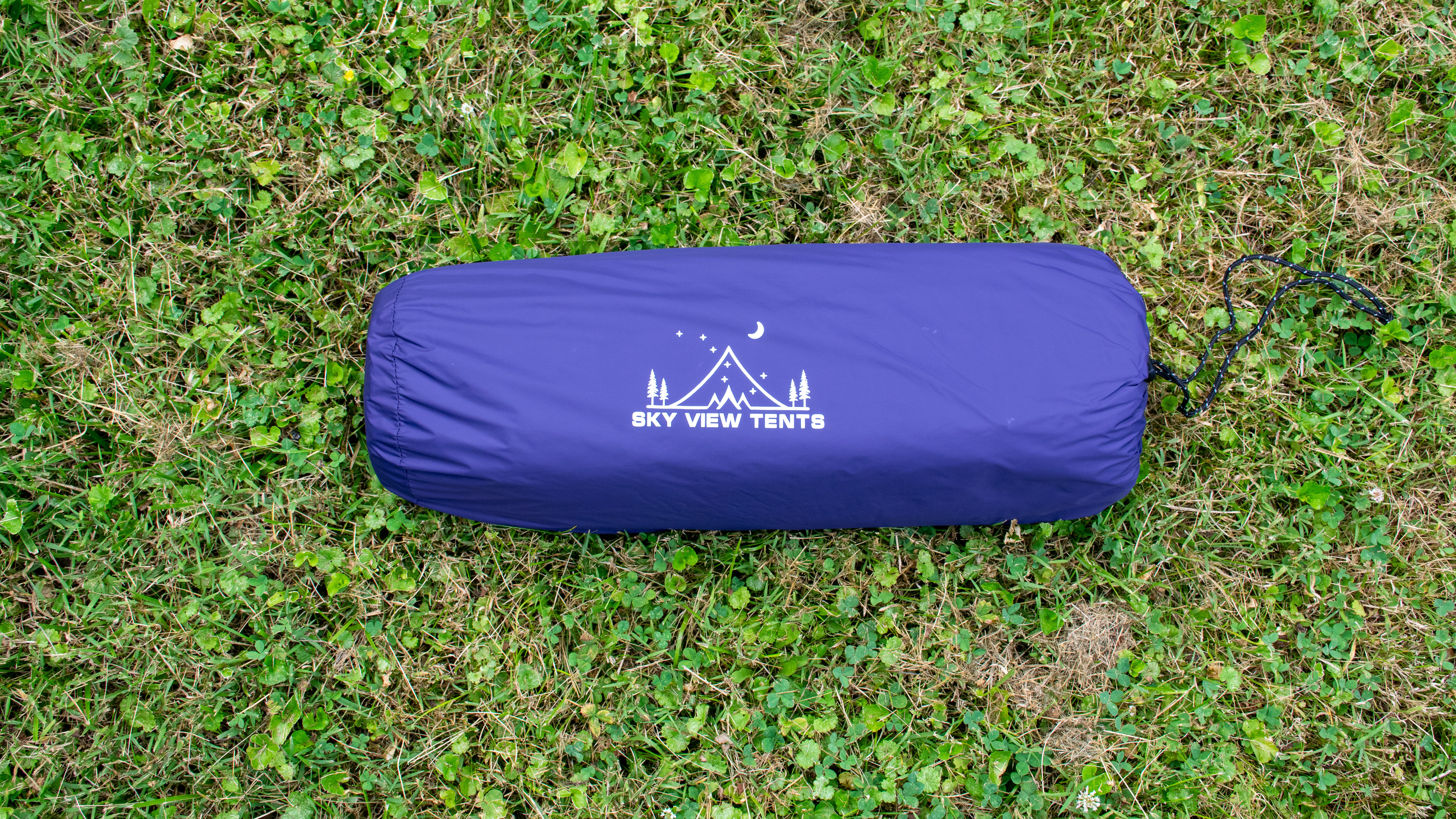 A purple nylon cylindrical bag rests on some grass.