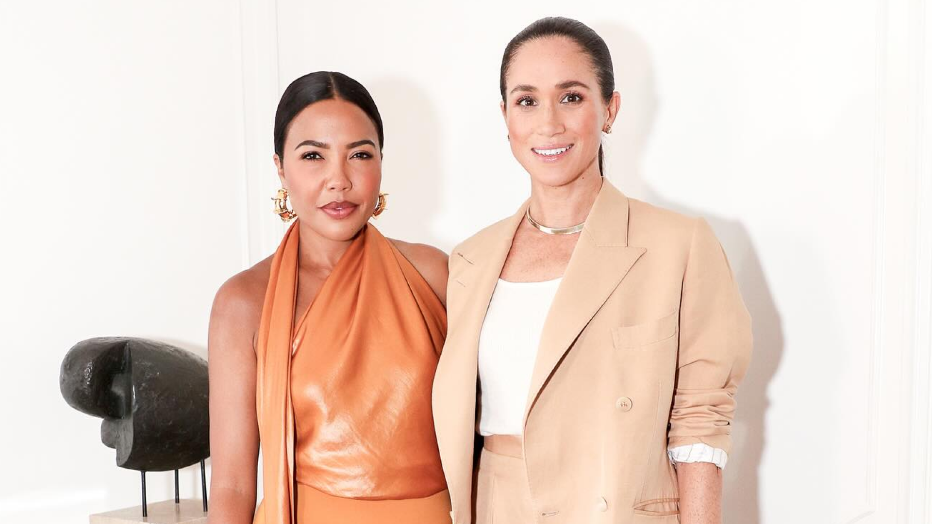 Meghan Markle wearing a beige suit standing with Emma Grede in an orange dress