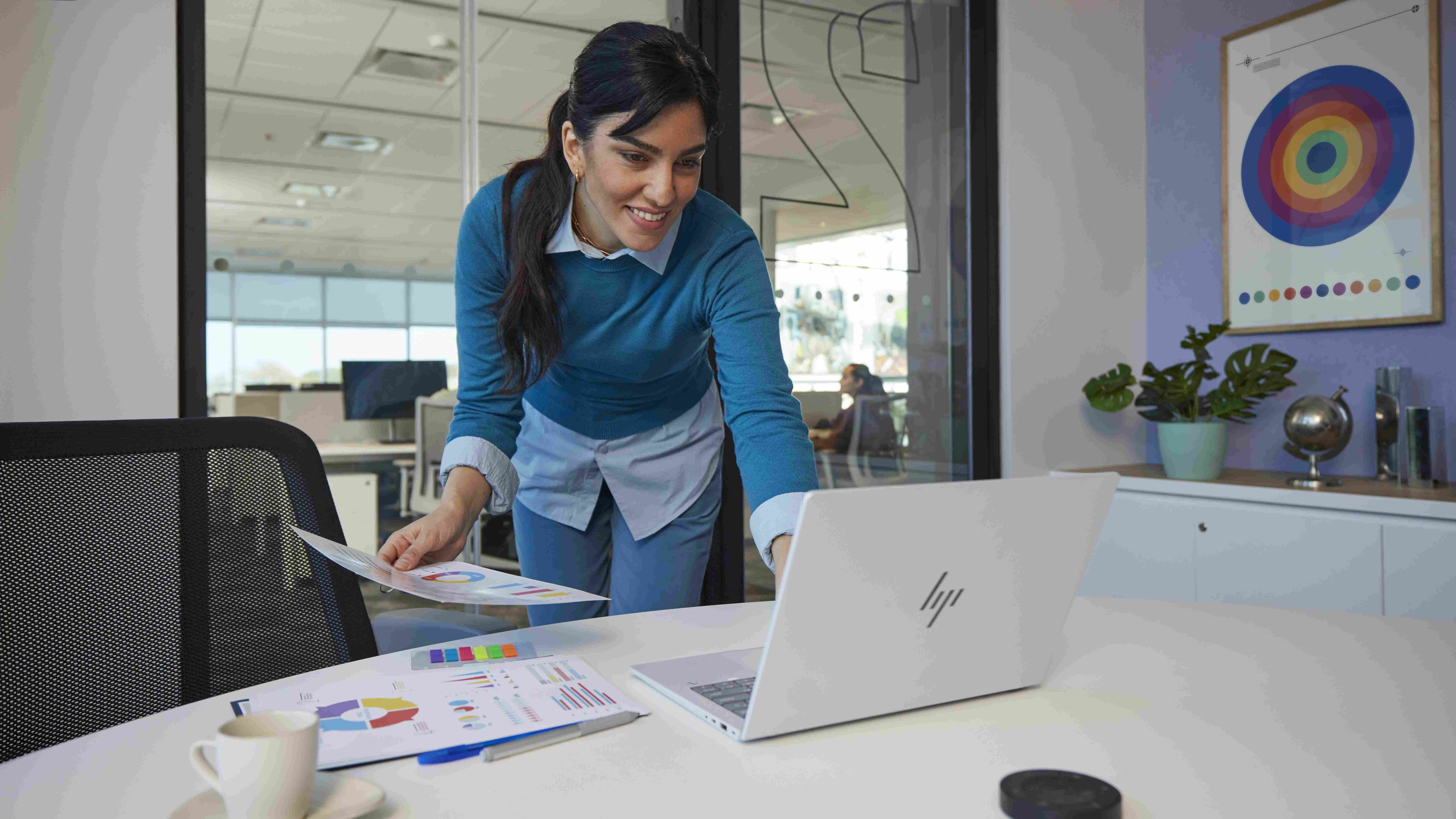 A woman bending down to use an HP EliteBook X 14 (G1i) on the table in front of her while holding papers.