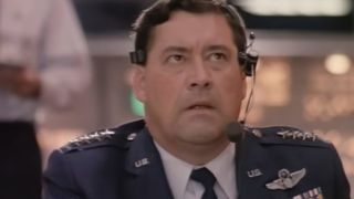 Barry Corbin with a telephone headset on and wearing a military uniform.