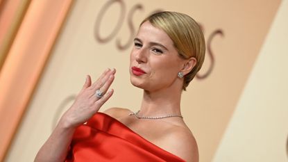 Jessie Buckley pictured on the Oscars red carpet in a red slink dress, holding her hand up with sheer, short nails and a large diamond ring