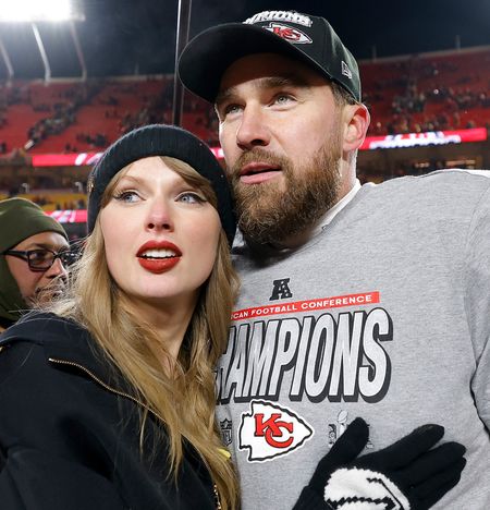 Taylor Swift wearing a black beanie and hoodie hugging Travis Kelce, who is wearing an AFC Champions shirt and black hat on the field after the 2025 AFC championship game