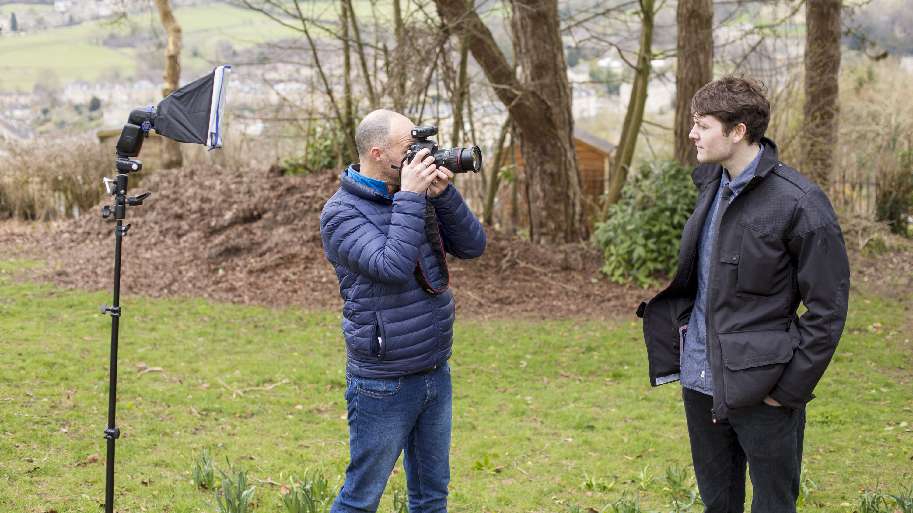 Bald man in blue jacket takes a photo of a white man with dark hair, shirt and tie and black jacket, grass and trees behind
