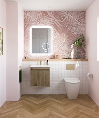 A small bathroom with pink and white leaf wallpaper on the walls, a rectangular mirror, a wooden vanity sink, white tiles and a toilet, and wooden herringbone flooring