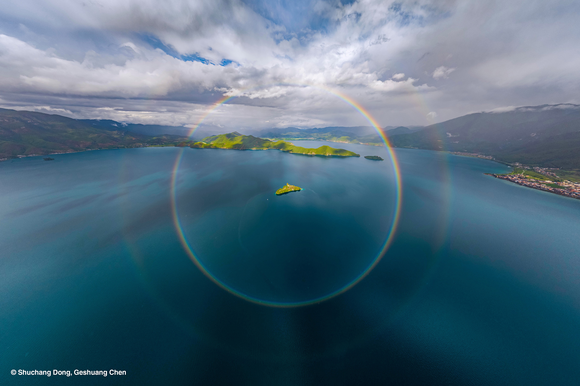 A stunning aerial view of a serene lake surrounded by green hills, featuring a vibrant rainbow halo overhead