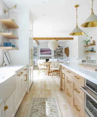A coastal kitchen with white cabinets and wooden wall shelves to the left, a white kitchen island with wooden drawers and an oven to the right, gold and gray pendant lights above it, and a wood floor with a runner rug