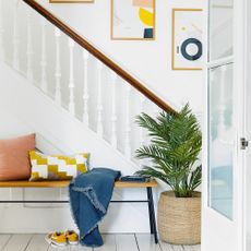 hallway decorating mistakes a white painted hallway and stairwell with a large plant in a basket a bench covered in cushions in graphic prints and similar graphic prints in frames up the stairs