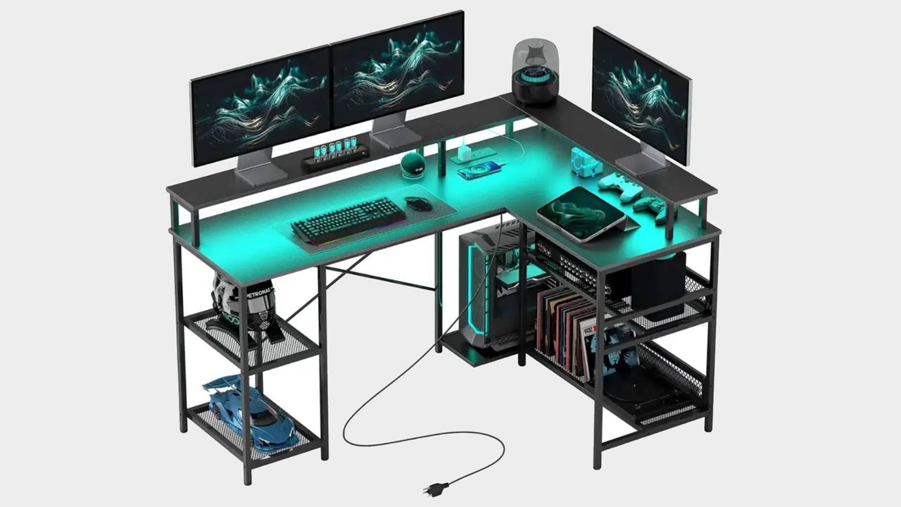 Homcom L-shaped desk with grey backdrop and blue RGB lights illumining surface with three monitors on top risers, controllers, keyboard, and tablet on surface, books on bottom shelves, and PC in back holder.
