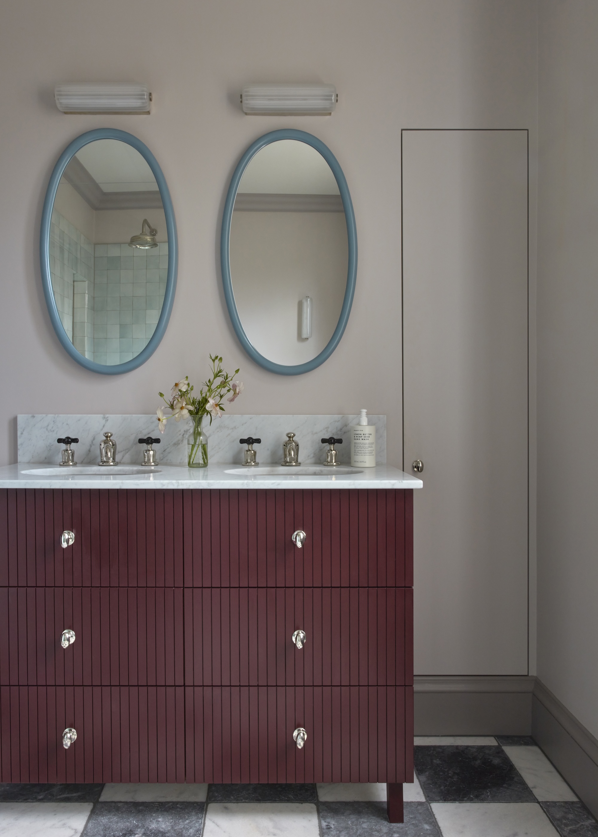 A guest bathroom with a maroon colored vanity with double basins. Above the vanity are two blue oval framed mirrors with lights above them.