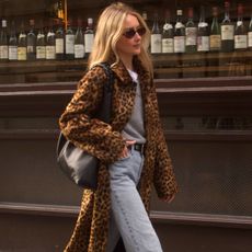 British style influencer Chloe Kath Butler walking on a London sidewalk wearing oval sunglasses, a leopard-print coat, gray V-neck sweater layered over a white T-shirt, studded black belt, black tote bag, and light-wash jeans