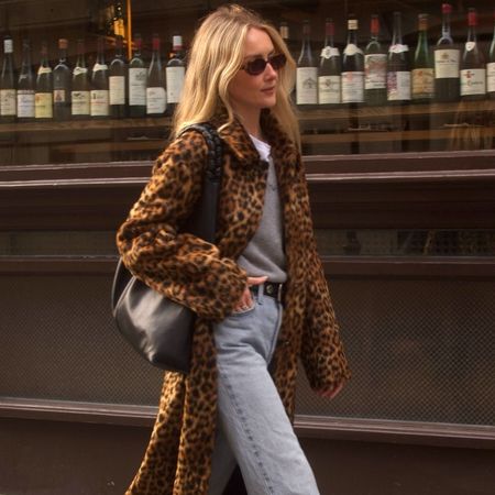 British style influencer Chloe Kath Butler walking on a London sidewalk wearing oval sunglasses, a leopard-print coat, gray V-neck sweater layered over a white T-shirt, studded black belt, black tote bag, and light-wash jeans
