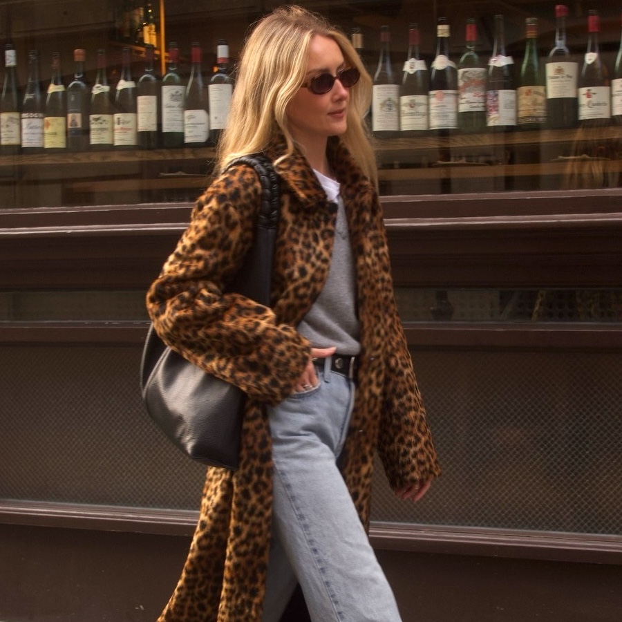 British style influencer Chloe Kath Butler walking on a London sidewalk wearing oval sunglasses, a leopard-print coat, gray V-neck sweater layered over a white T-shirt, studded black belt, black tote bag, and light-wash jeans