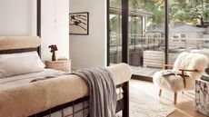 A white bedroom with a fuzzy chair, a stone side table, a wood bedside table with a vase of flowers and a pendant lamp above, and a bed with upholstered head and foot boards