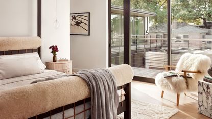 A white bedroom with a fuzzy chair, a stone side table, a wood bedside table with a vase of flowers and a pendant lamp above, and a bed with upholstered head and foot boards