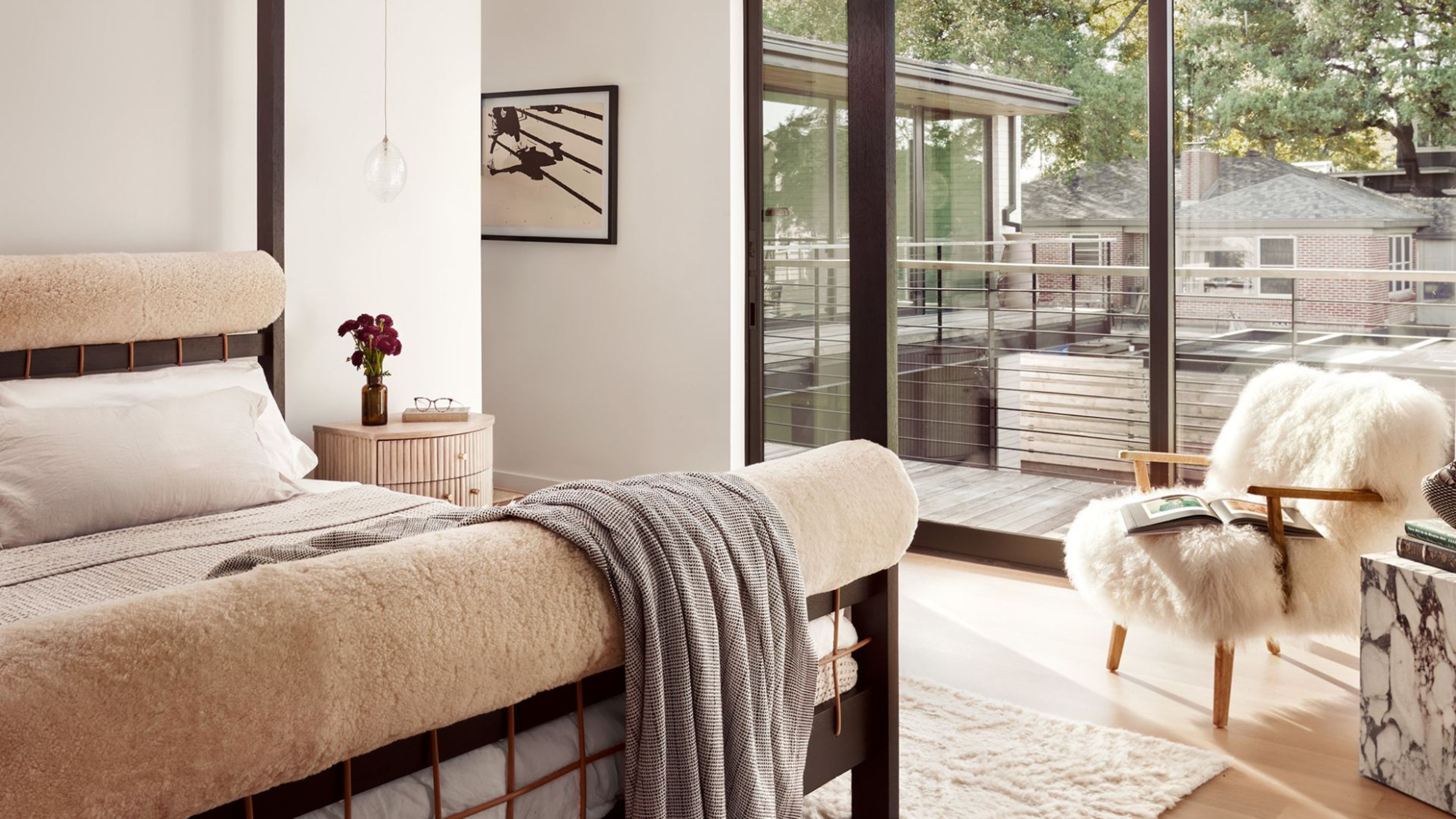 A white bedroom with a fuzzy chair, a stone side table, a wood bedside table with a vase of flowers and a pendant lamp above, and a bed with upholstered head and foot boards