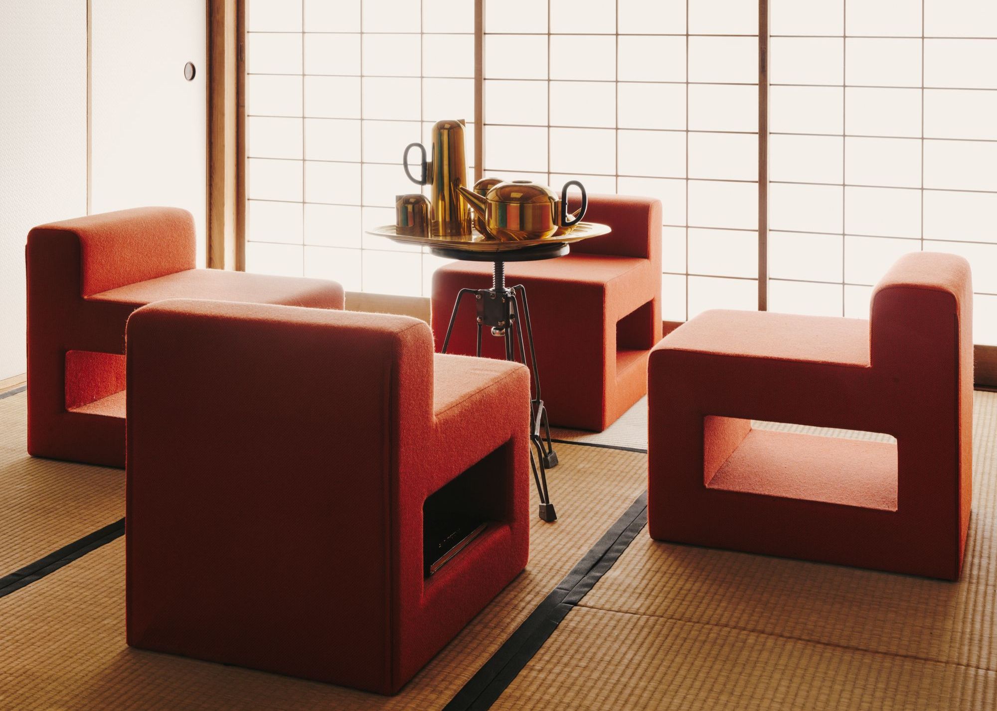 A Japanese tea room with shoji screens, tatami mats and angular yet soft burnt orange dining chairs with a gold tea set on a round table in the center