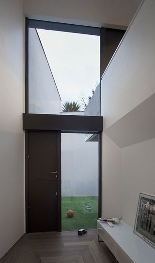 A corridor with high white ceiling and closed black door with a side & top glass window, leading to a courtyard/play area with green grass with a basket ball and 2 rackets on the grass. On the right of the corridow is a low white cabinet with a aeroplane toy and picture frame on it.