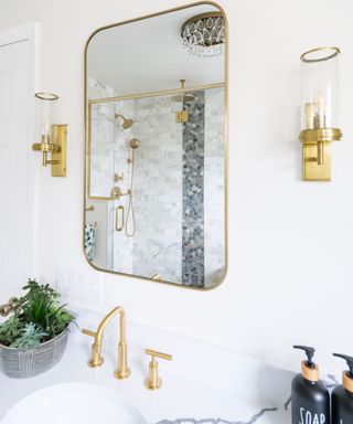 A bathroom with a curved gold mirror, two gold light wall sconces, a white sink with gold taps, a green plant to the left and two black soap dispensers to the right