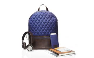 A backpack with a blue diamond pattern and a brown leather zip pocket next to headphones, books and a cellphone in a blue holder.