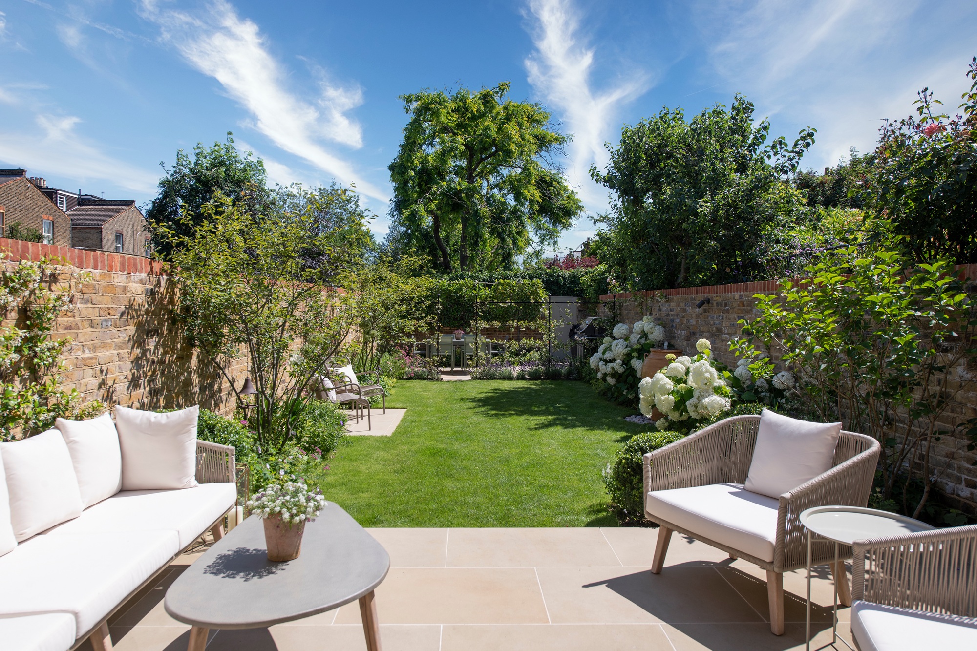 a modern garden with a patio, a large lawn, and multiple seating areas, surrounded by trees