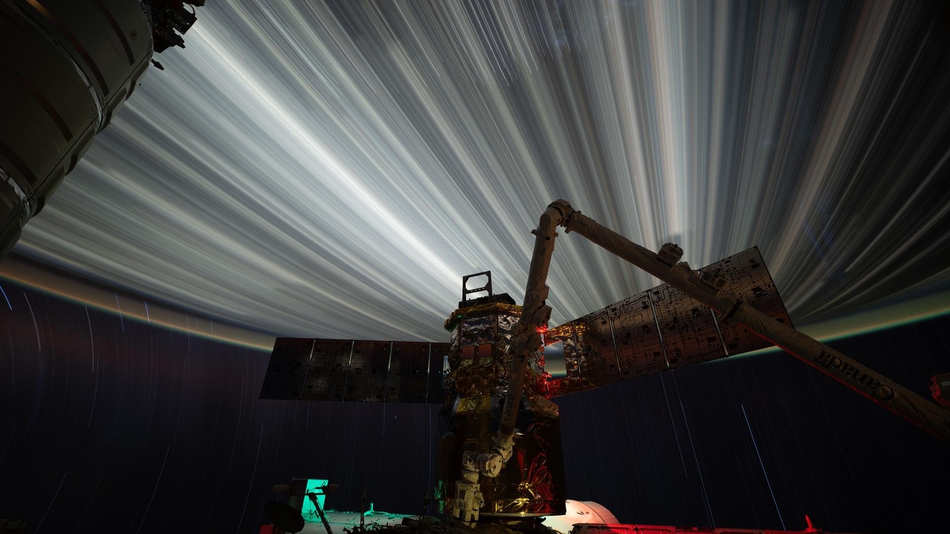 a half-circle made of streaks of bright light dominates the top half of the image as the silhouette of a robotic arm and solar panels can be seen below