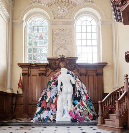 Inside Blenheim Palace looking at a white naked statue with its back to the photographer. In front of the statue is a pile of clothes at the same height as the statue. 