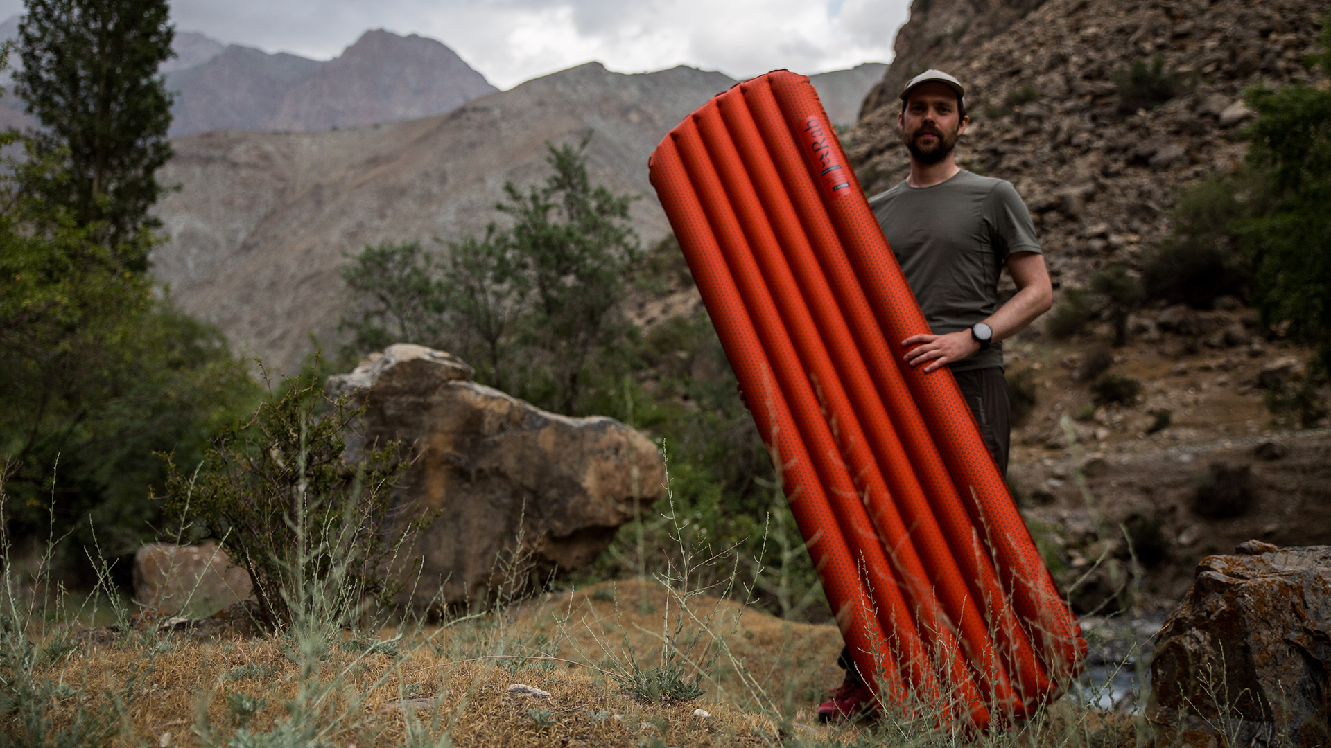 A man stands in rocky countryside holding an orange Rab Ionosphere 5.5 Sleep Mat