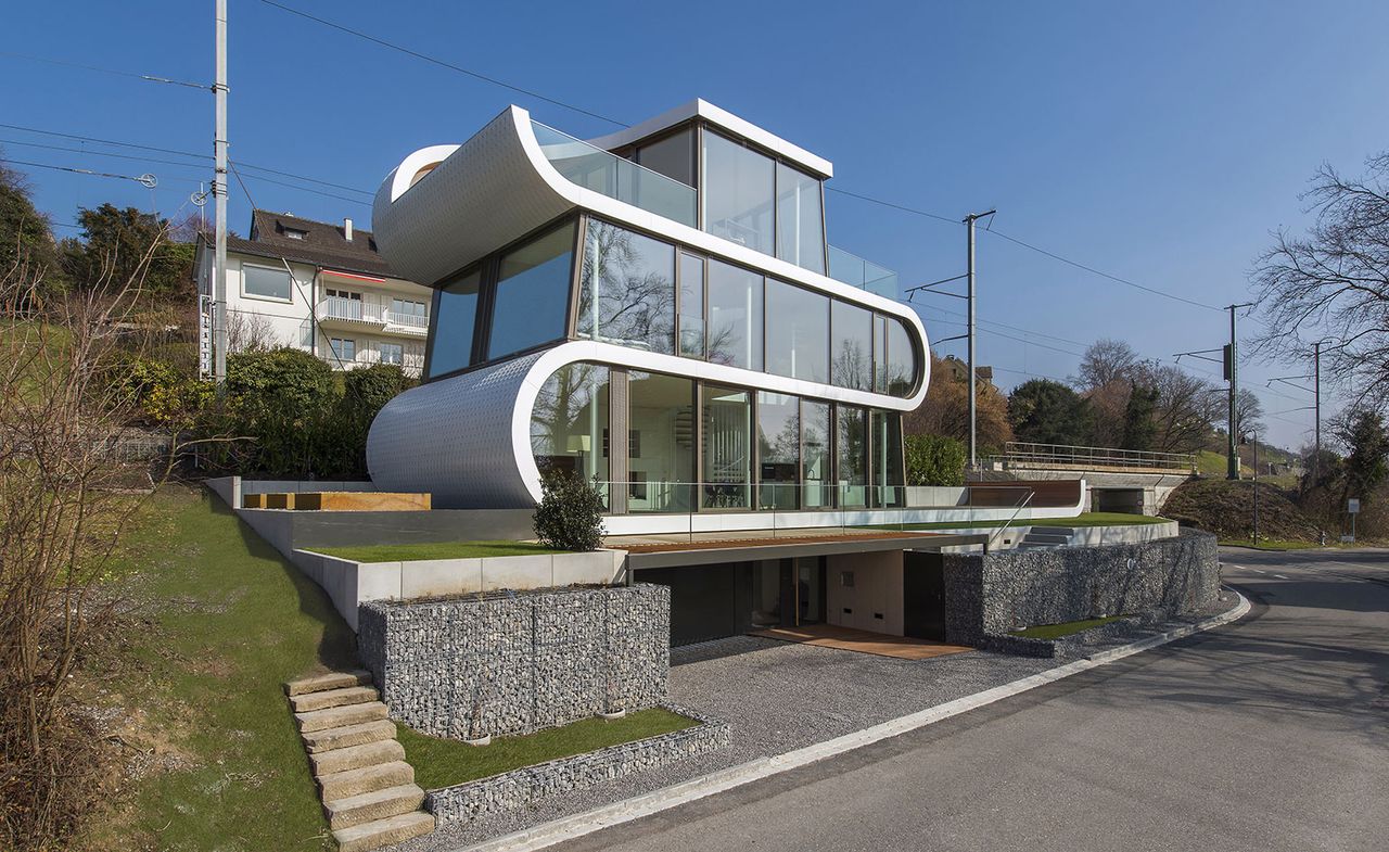 The exterior of an elevated curved structural black and grey residential building with floor to ceiling doors and windows. stair case on the right and left of the building. Building of off a road