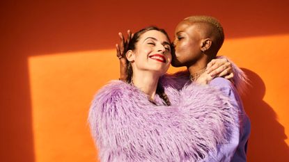 A couple in an embrace and wearing bright purple clothing standing infront of a bright orange wall