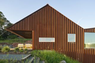 Close up exterior view of the Corten steel facade of Triple Barn house during the day. The yellow and white outdoor furniture and surrounding greenery can also be seen