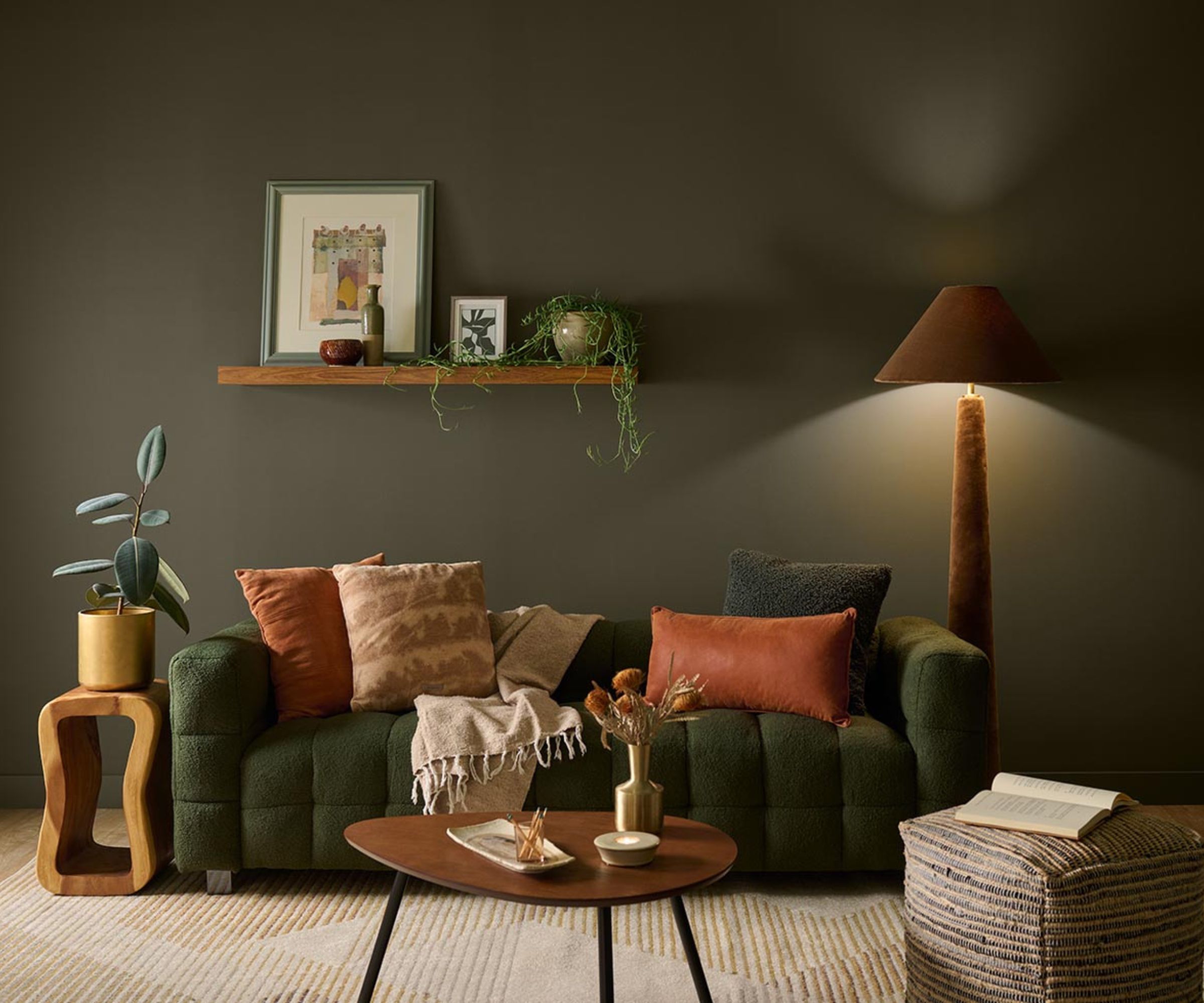 Living room painted olive green, a green quilted couch decorated with orange and green pillows, a midcentury coffee table, a pouffe, and a brown floor lamp in the corner.