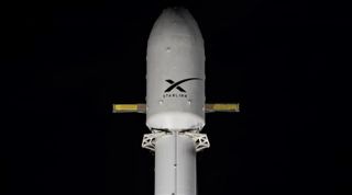 A white rocket topped with a payload fairing stands on the launch pad in a close-up set against a black sky.