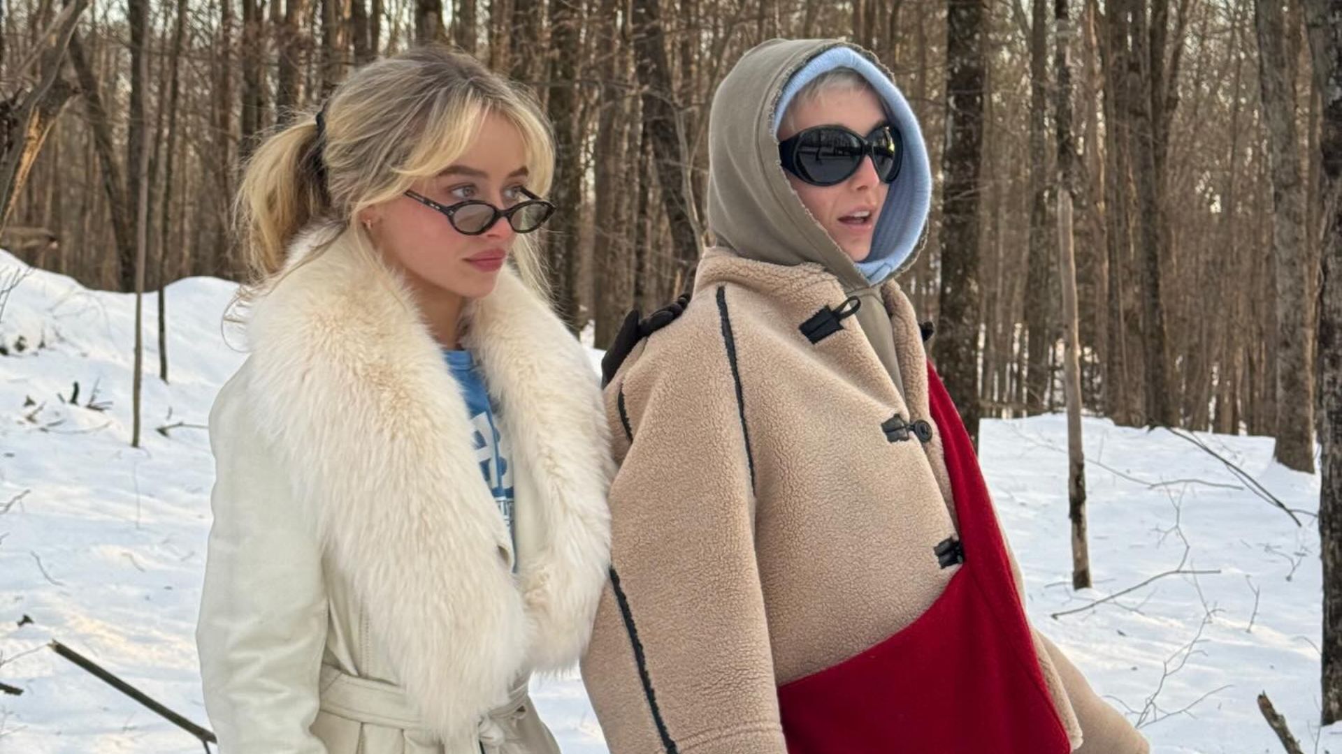 Sabrina Carpenter wears a white fur-trimmed coat over a blue t-shirt, standing next to Emma Chamberlin who wears a borge jacket with a khaki hood.
