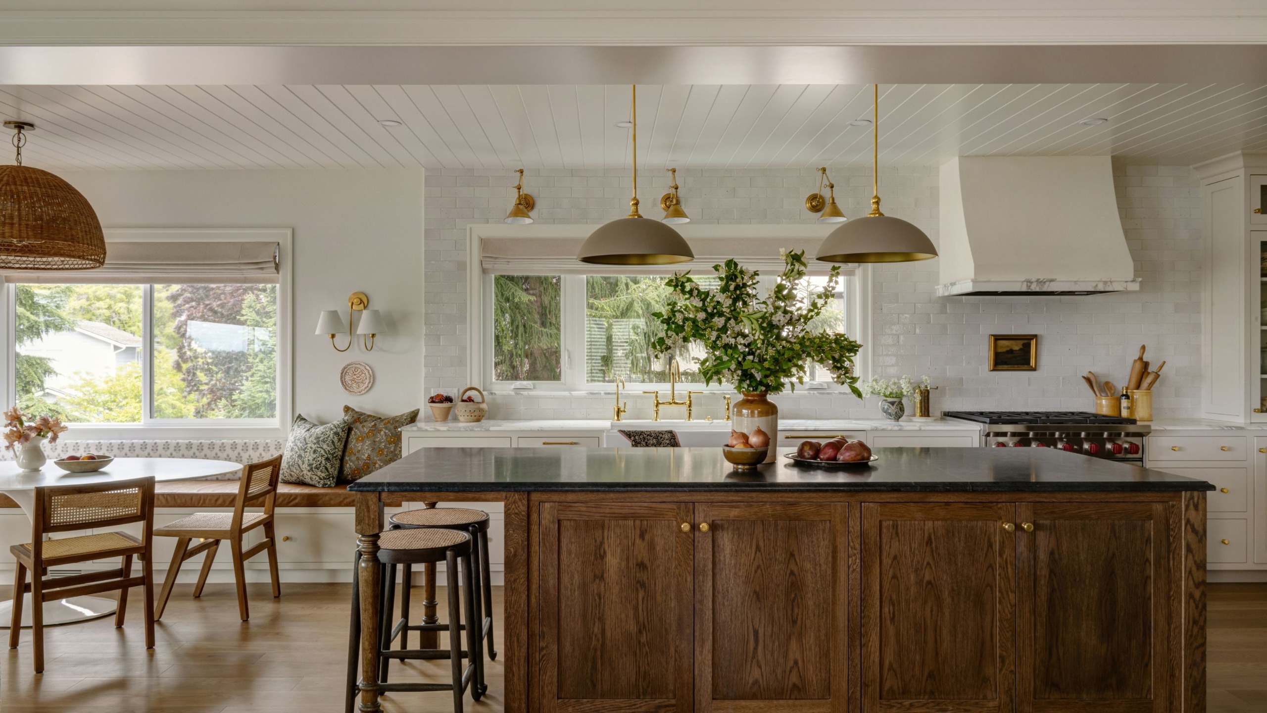 A large kitchen with transitional decor. White walls, wood flooring, and a dark wood kitchen island with soapstone countertops. Off-white backsplash tiles, white marble counters, and warm white cabinets.