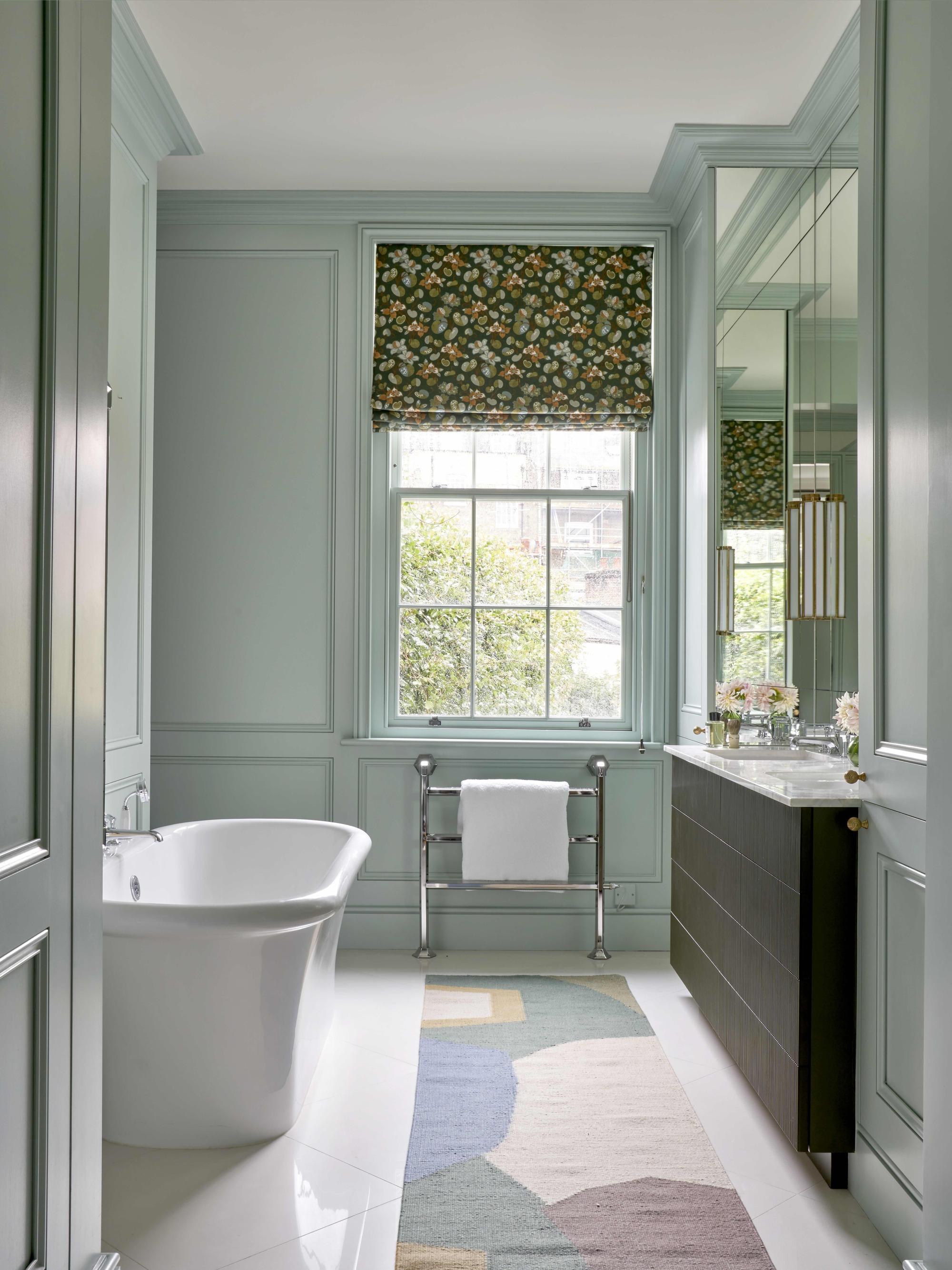 a light minty blue bathroom with freestanding bath, heated towel rail, dark vanity with white counter, mirrored walls, a window with a patterned roman blind, white glossy floor tiles and a colorful bathroom rug