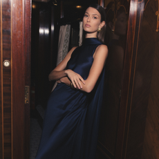 Model wearing a dark blue satin Reiss dress and gold Reiss earrings stood in a dark wooden doorway for Reiss discount codes