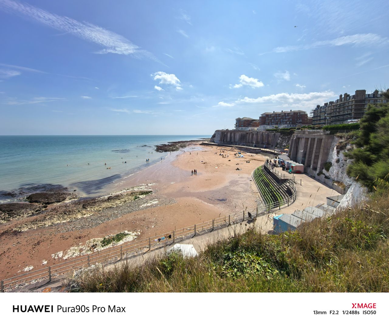 Wide coastal view of a sandy beach beneath chalk cliffs and seafront buildings on a bright sunny day.