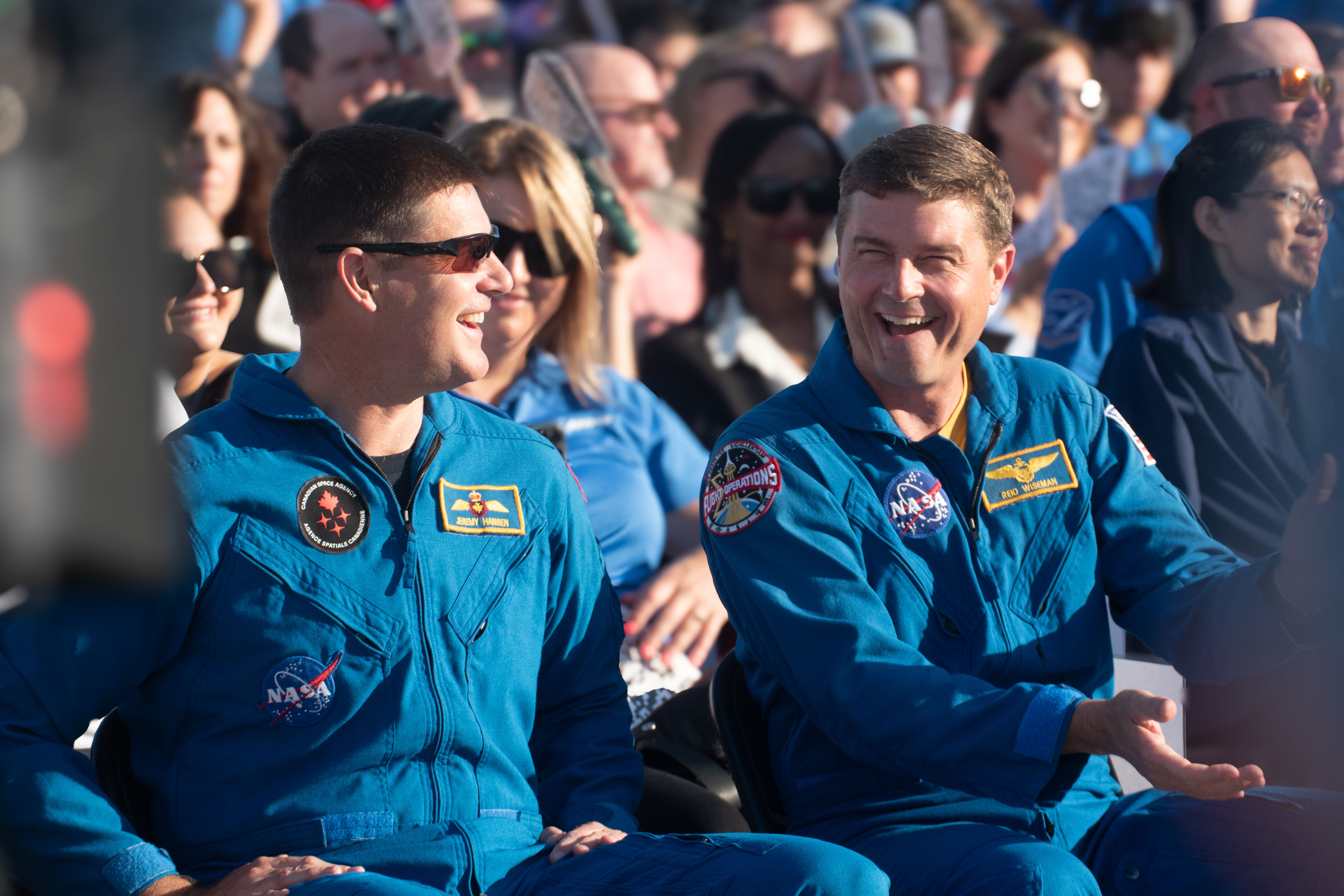 Two men in blue jumpsuits laughing while sitting in a crowd.