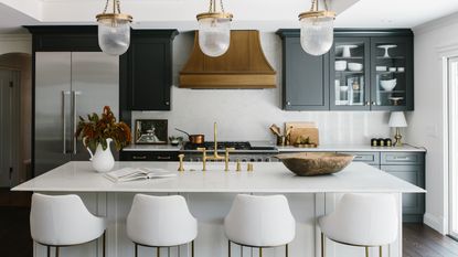 A kitchen with white marble topped island, white upholstered bar stools, black kitchen cabinets, bronze hood and pendant lights