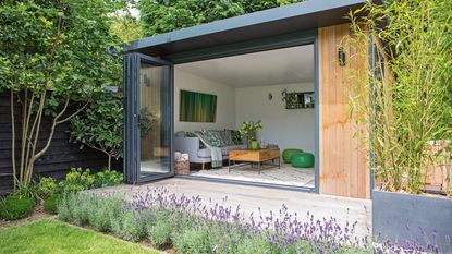 A garden summerhouse with sliding glass door, white walls, and green decorative accents, surrounded by plants