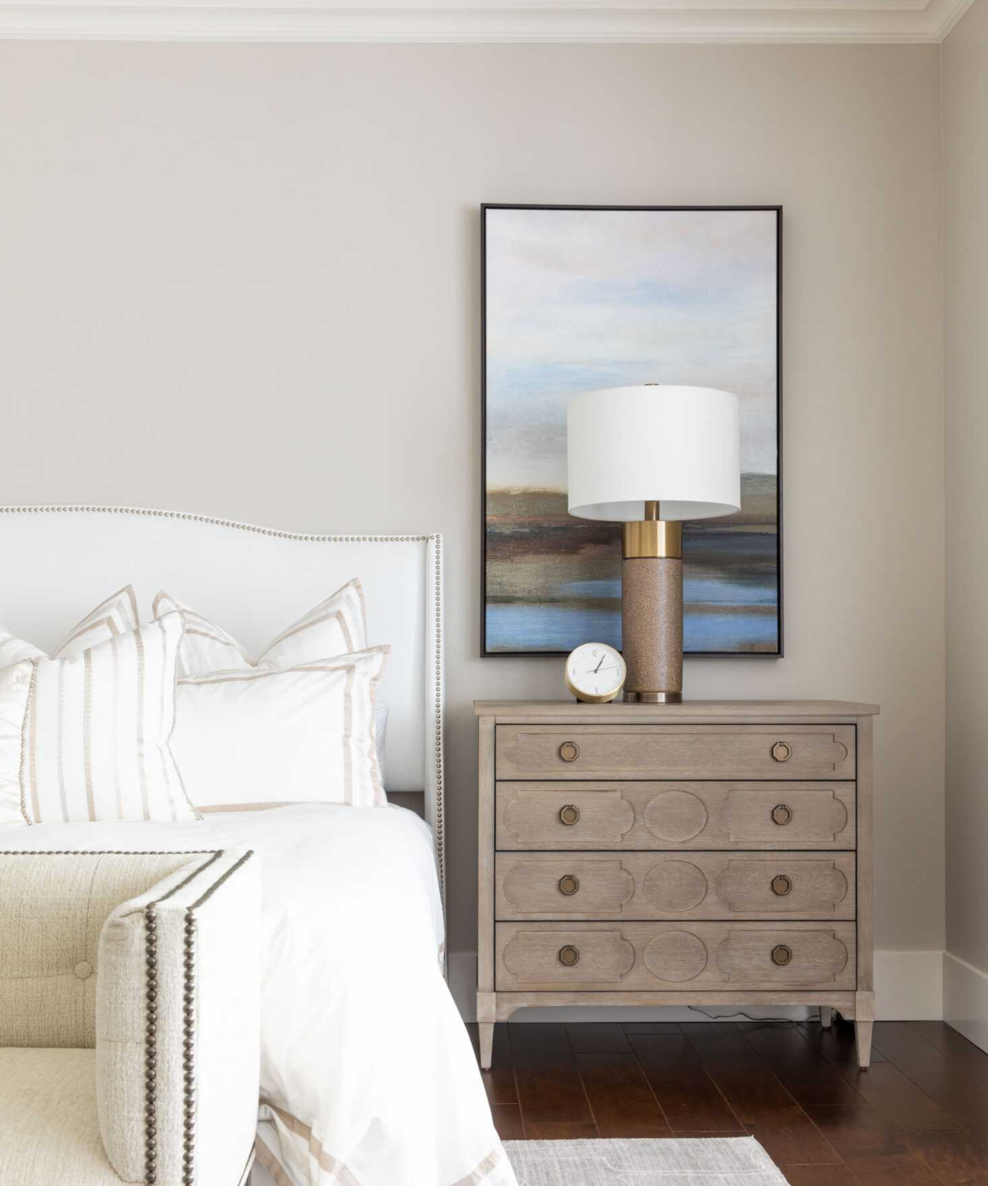 A beige bedroom with a white bedframe, a scenic painting, and a wooden side table