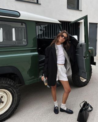 influencer mode wanita Emilie T. berpose di luar Land Rover hijau mengenakan kacamata hitam persegi berwarna cokelat, blazer hitam besar yang dilapisi di atas kemeja kancing bergaris terbuka dan kaos pointelle putih, celana boxer katun putih, kaus kaki putih, dan sepatu pantofel hitam.