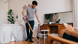Man with dark brown hair wearing a grey tshirt vacuuming floor in bedroom.