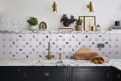 How to install a backsplash, shown in a white kitchen with Delft tiles, a white marble countertop and shelf, black cabinetry and antique-style gold faucet and wall lighting.
